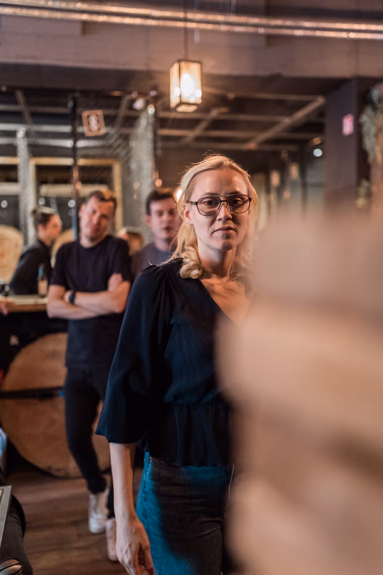 Blonde Frau mit Brille in einer Bar, im Hintergrund wartende Kollegen beim Firmenausflug